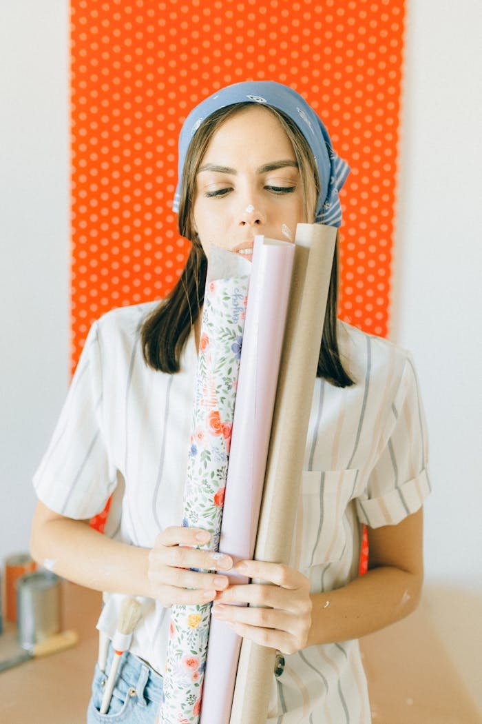 about-bg Woman with bandana holding various wallpaper rolls indoors, planning home decor.